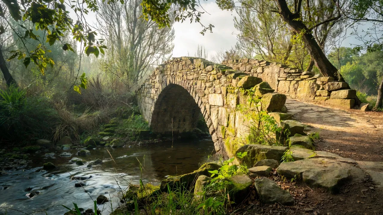 An old stone arch bridge over a small stream, moss-covered, in dappled morning light.